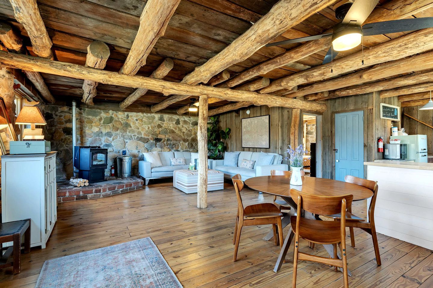 Un intérieur rustique avec un mur en pierre, des poutres en bois au plafond, une cheminée en pierre, un canapé, une table à manger et un coin cuisine.