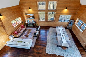 A spacious room with a wooden floor, a sofa set, a dining table with chairs, and large windows offering a view of the outdoors.