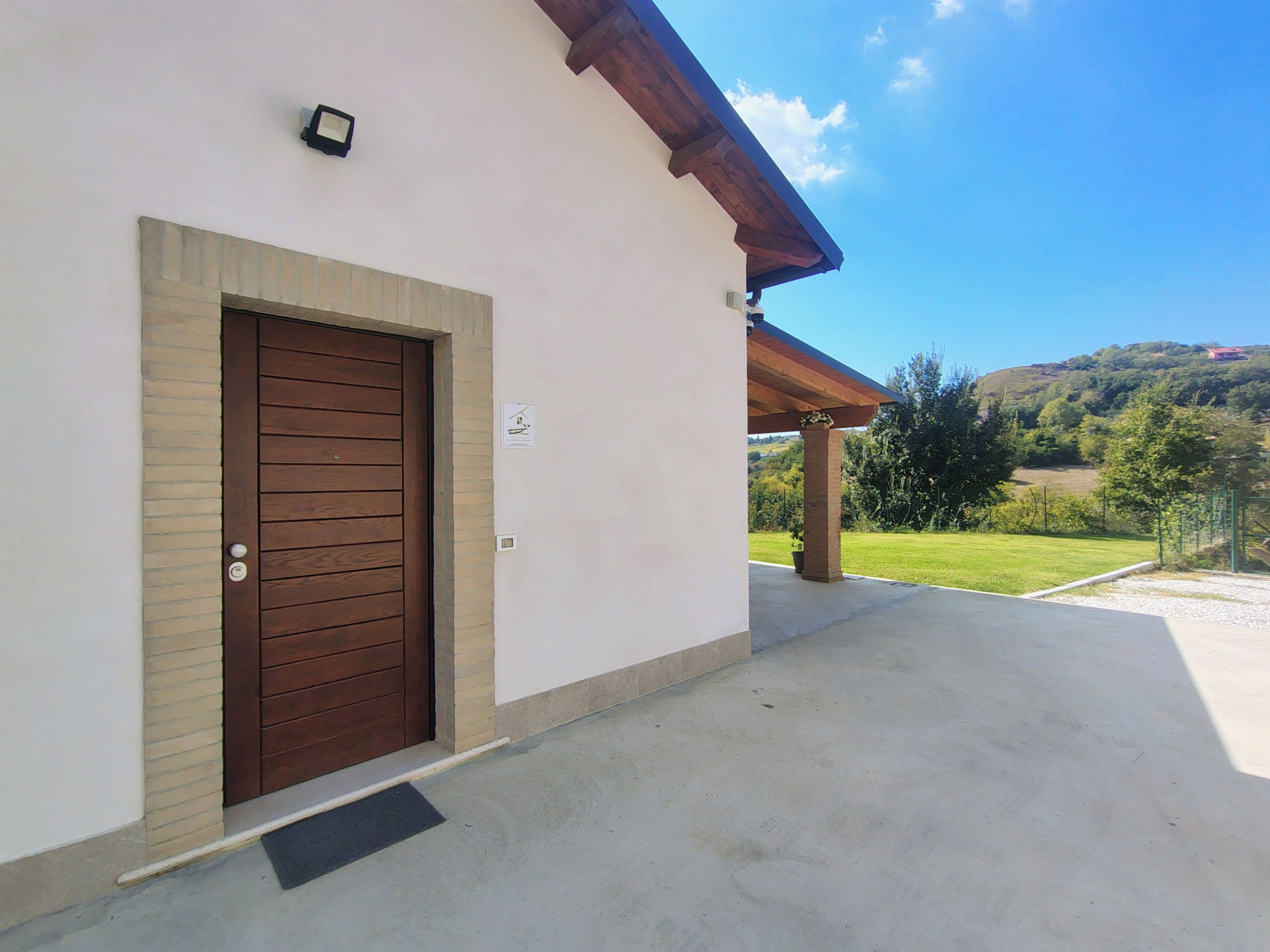 Un edificio con una porta marrone, un piccolo cartello sopra la porta e un'area esterna coperta con vista su un paesaggio verde.