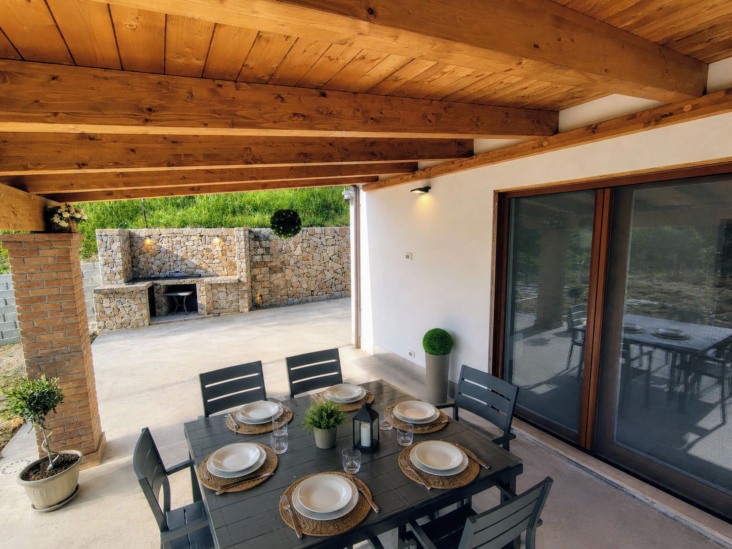 Un patio exterior con una barbacoa de piedra, una mesa de comedor preparada para cuatro personas y un amplio ventanal corredero.
