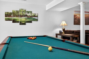A pool table with balls, a cue stick, and a lamp. There are three framed pictures on the wall.