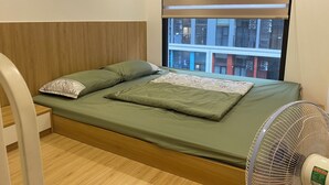 A single bed with a wooden headboard, a green bedspread, and two pillows. A standing fan is positioned to the right of the bed.