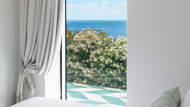 A bedroom with a large window, a bed, and a view of the sea and flowers. - Estate4home - Villa Nerea (Massa Lubrense)