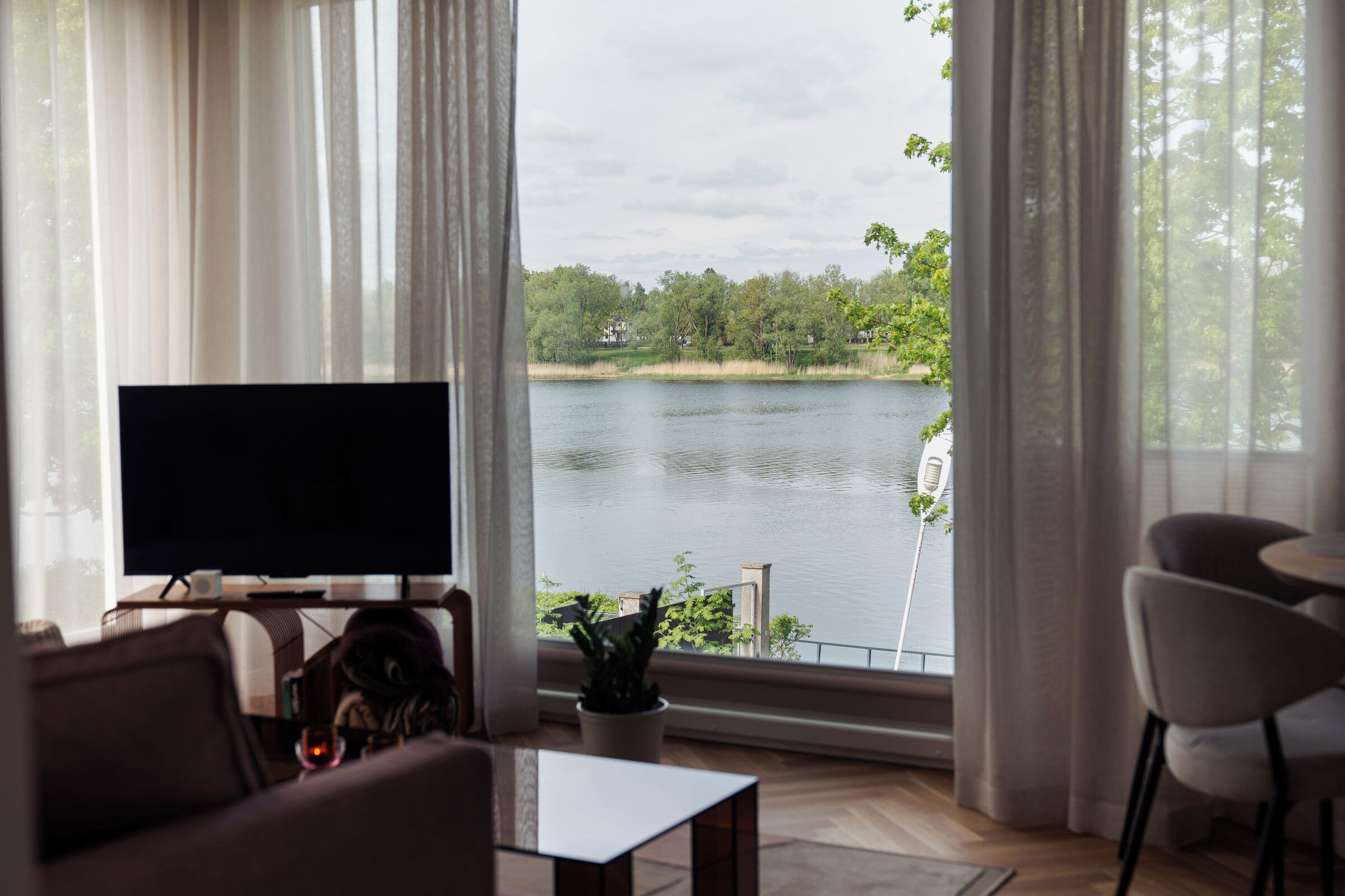 A living room with a TV, a coffee table, and a view of a lake through large windows.