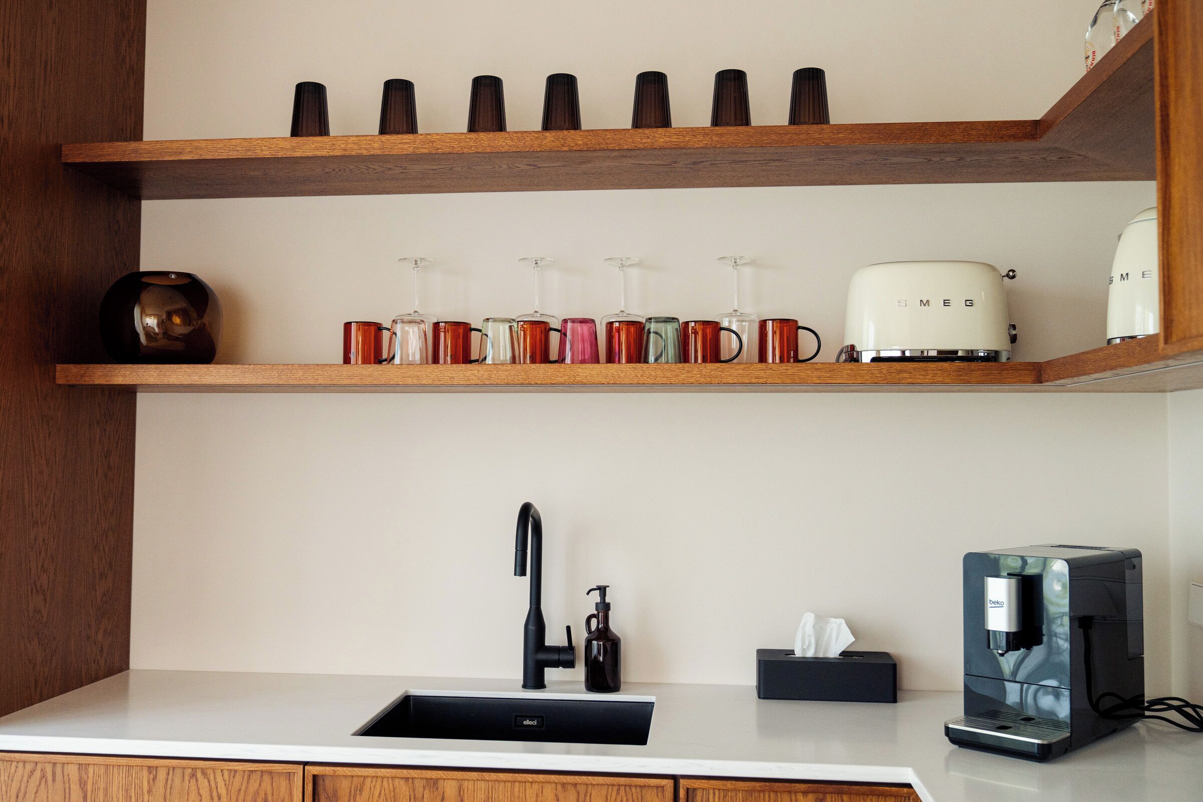 A kitchen with a white countertop, a black faucet, a coffee machine, and a shelf with various mugs and a vase.