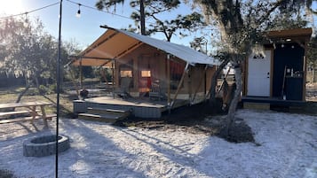 Un espace extérieur abrité par une tente, comprenant une table de pique-nique, un foyer et un petit bâtiment doté d’une porte.