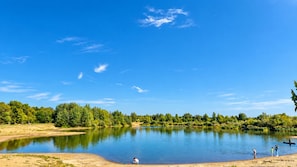 Un lac paisible aux rives sablonneuses, entouré d’une végétation luxuriante et d’arbres, sous un ciel bleu dégagé.