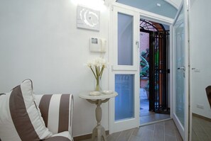 A room with a striped chair, a small table with a vase of flowers, a door leading outside, and a wall-mounted air conditioning unit. - Colosseo & Fori Imperiali Apartment (Rome)