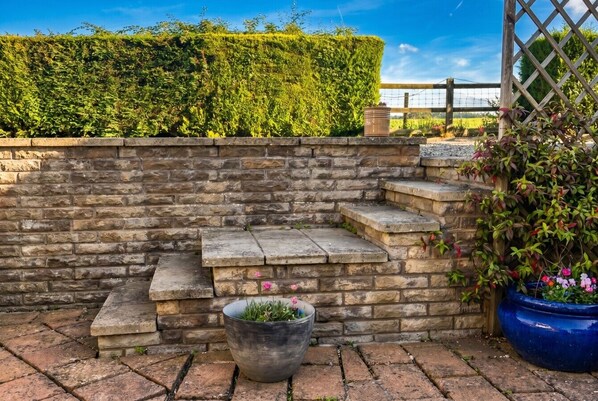 A stone wall with steps leading up to a wooden fence, a potted plant with pink flowers, and a blue pot with greenery.
