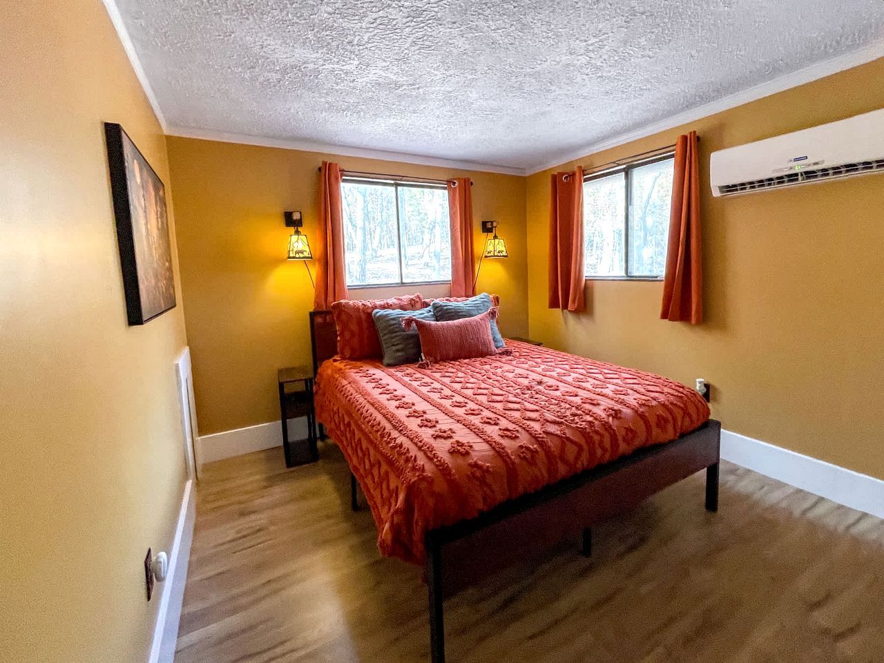 A bedroom with a bed, two windows with curtains, an air conditioner, and a framed picture on the wall.