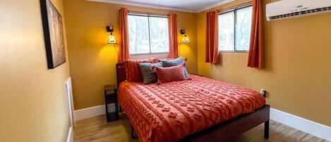 Ein Schlafzimmer mit einem Bett, zwei mit Vorhängen versehenen Fenstern, einer Klimaanlage und einem gerahmten Bild an der Wand.