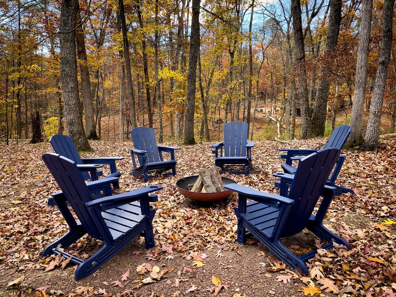 A group of Adirondack chairs arranged around a fire pit in a forested area.
