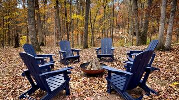 A group of Adirondack chairs arranged around a fire pit in a forested area.