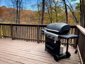 Ein Grill auf einer Holzterrasse mit Geländer, umgeben von Bäumen mit Herbstlaub.