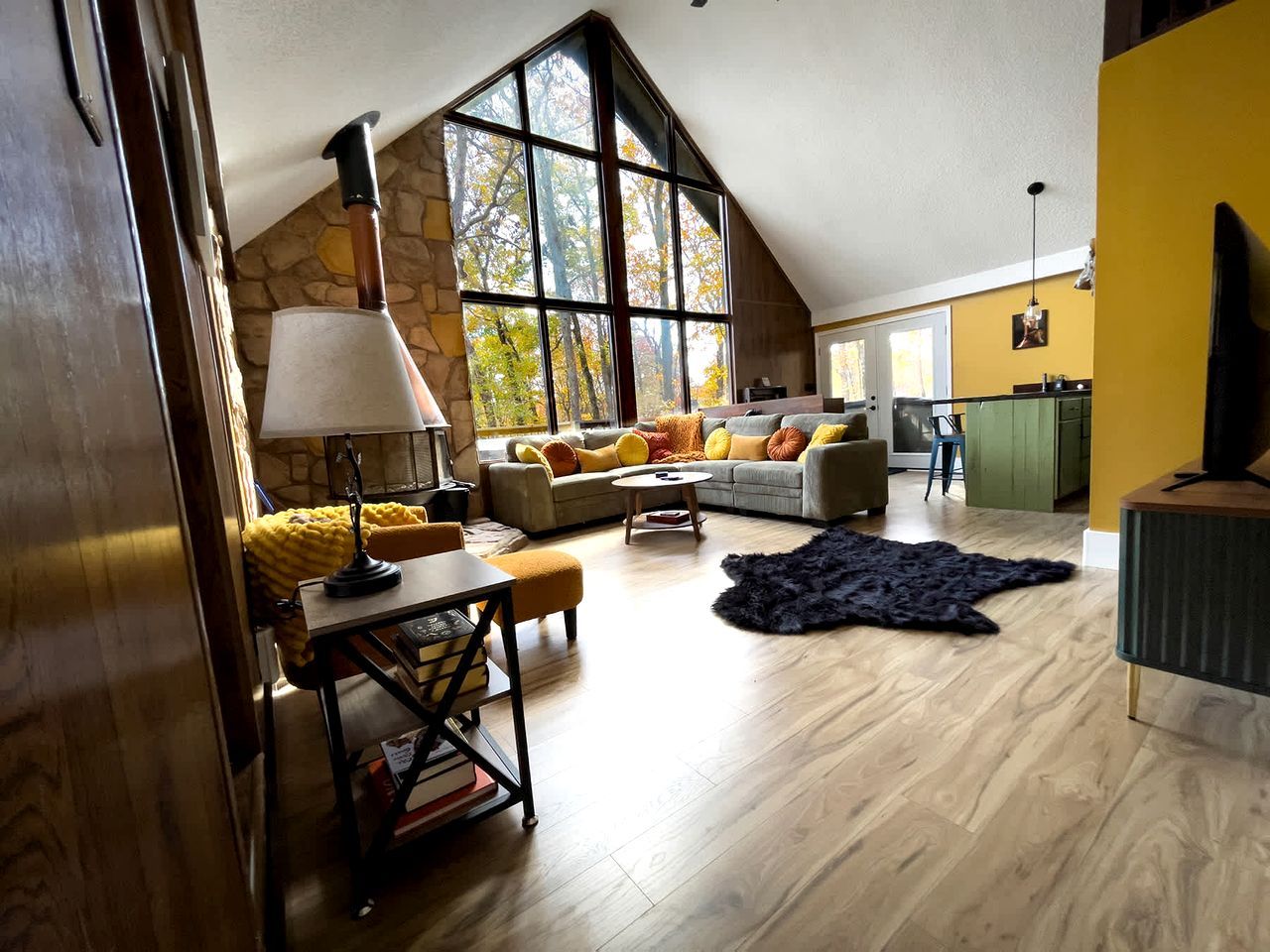 A spacious loft with a high ceiling, a stone wall, a large window, a sofa set, a dining table, and a television.