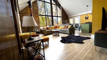 A spacious loft with a high ceiling, a stone wall, a large window, a sofa set, a dining table, and a television.