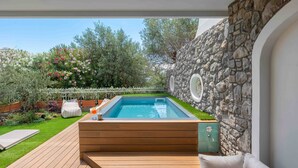Una piscina amb un solàrium de fusta i zona de seients, envoltada per una paret de pedra i vegetació abundant.