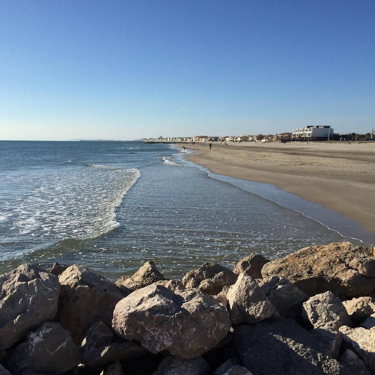 Une plage avec un premier plan rocheux, une rive sablonneuse et des bâtiments en arrière-plan.
