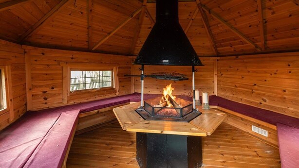 Interior d'una cabana de fusta amb una xemeneia central, parets de fusta i un sostre a dues aigües.
