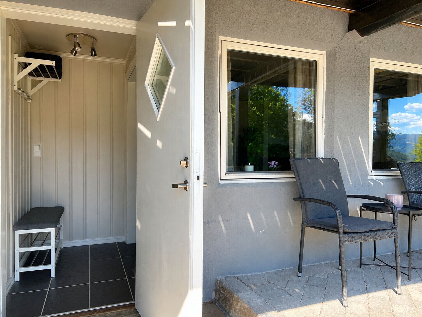 Een kamer met een witte deur, een klein opstapje naar een betegelde vloer, een raam met uitzicht op groen, en een stoel ernaast.