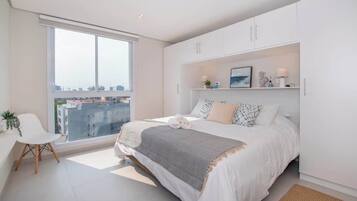 Un dormitorio con un ventanal grande, una cama, una silla y un armario blanco.