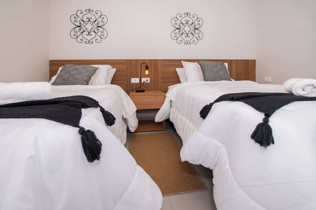 Habitació d'hotel amb dos llits, un capçal de fusta, una tauleta de nit amb una làmpada i una obra d'art decorativa a la paret.
