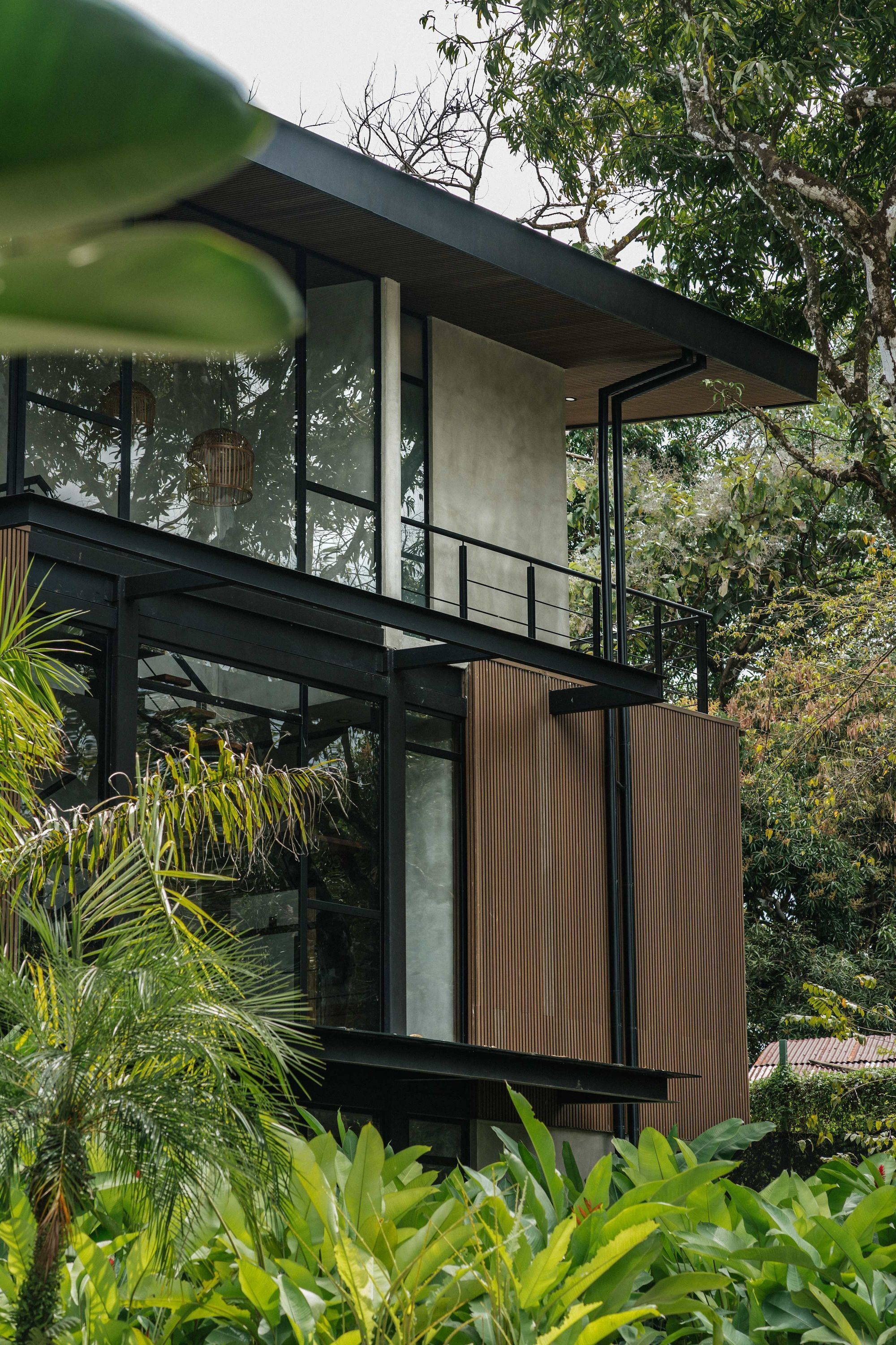A modern two-story building with large windows, a balcony, and surrounding lush greenery.
