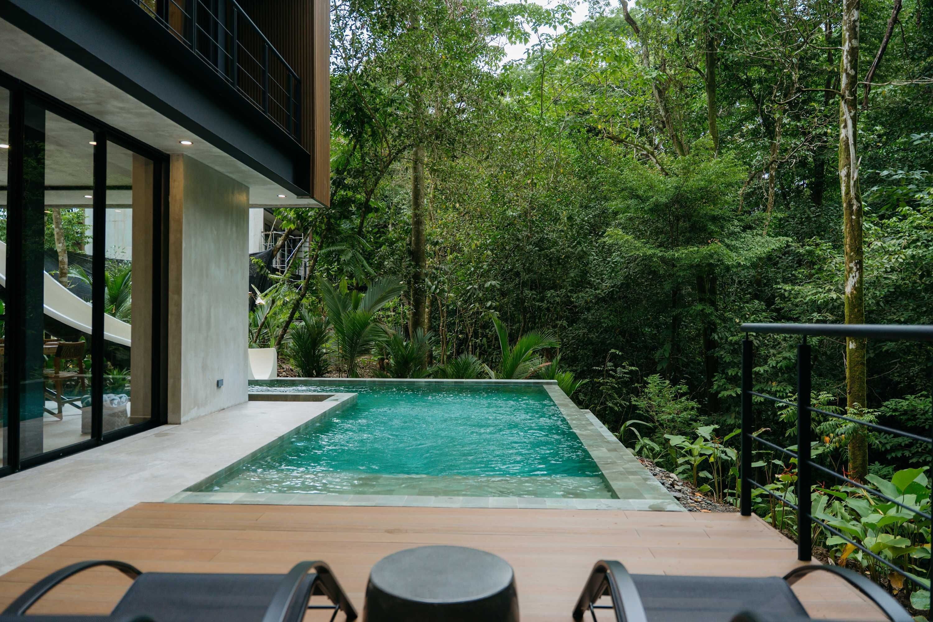A modern outdoor pool area with a wooden deck, lounge chairs, and a staircase leading to a building.