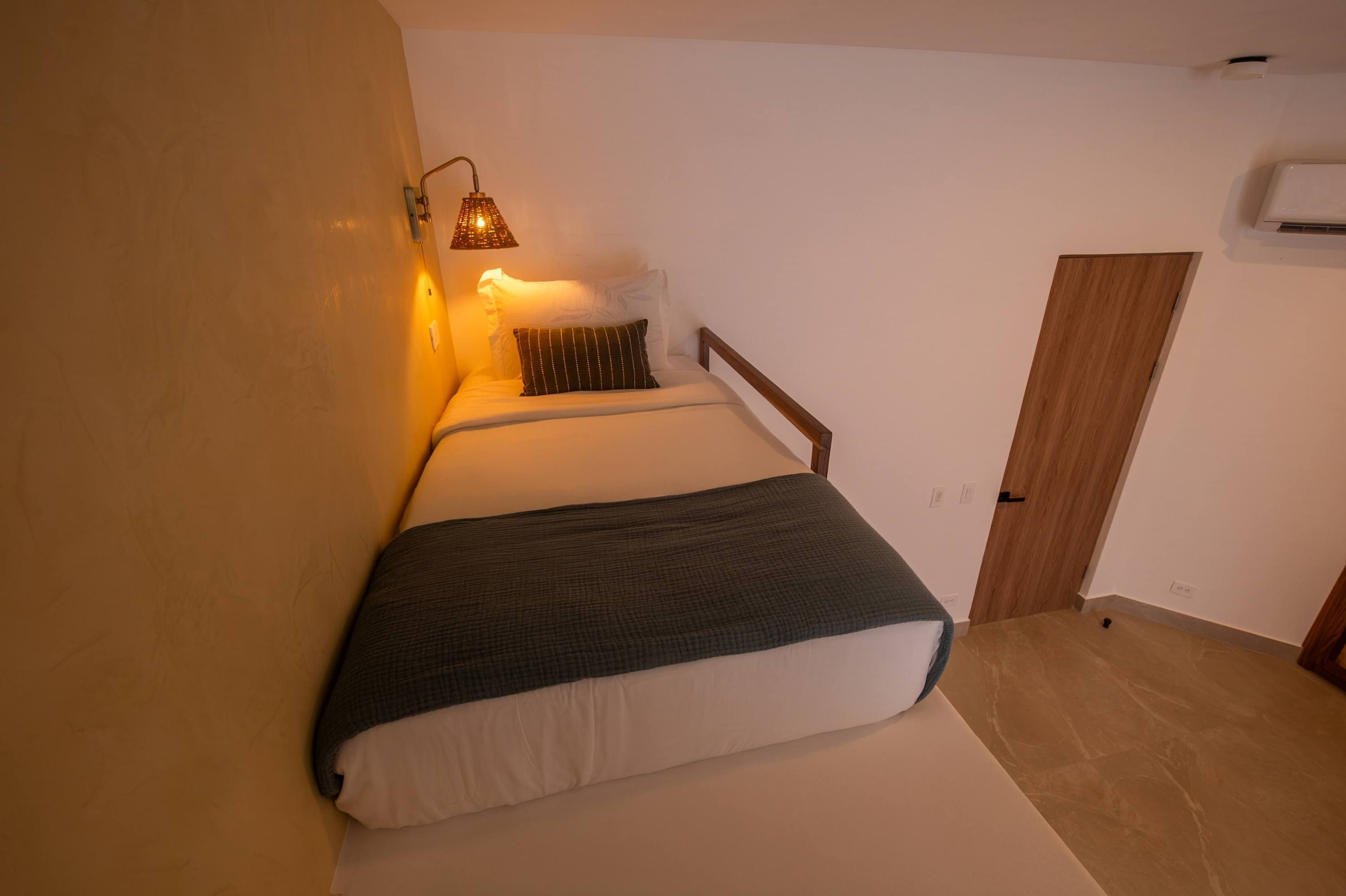 Une petite chambre à coucher, chaleureuse et accueillante, avec un lit, une lampe de chevet, une porte en bois et un appareil de climatisation fixé au mur.