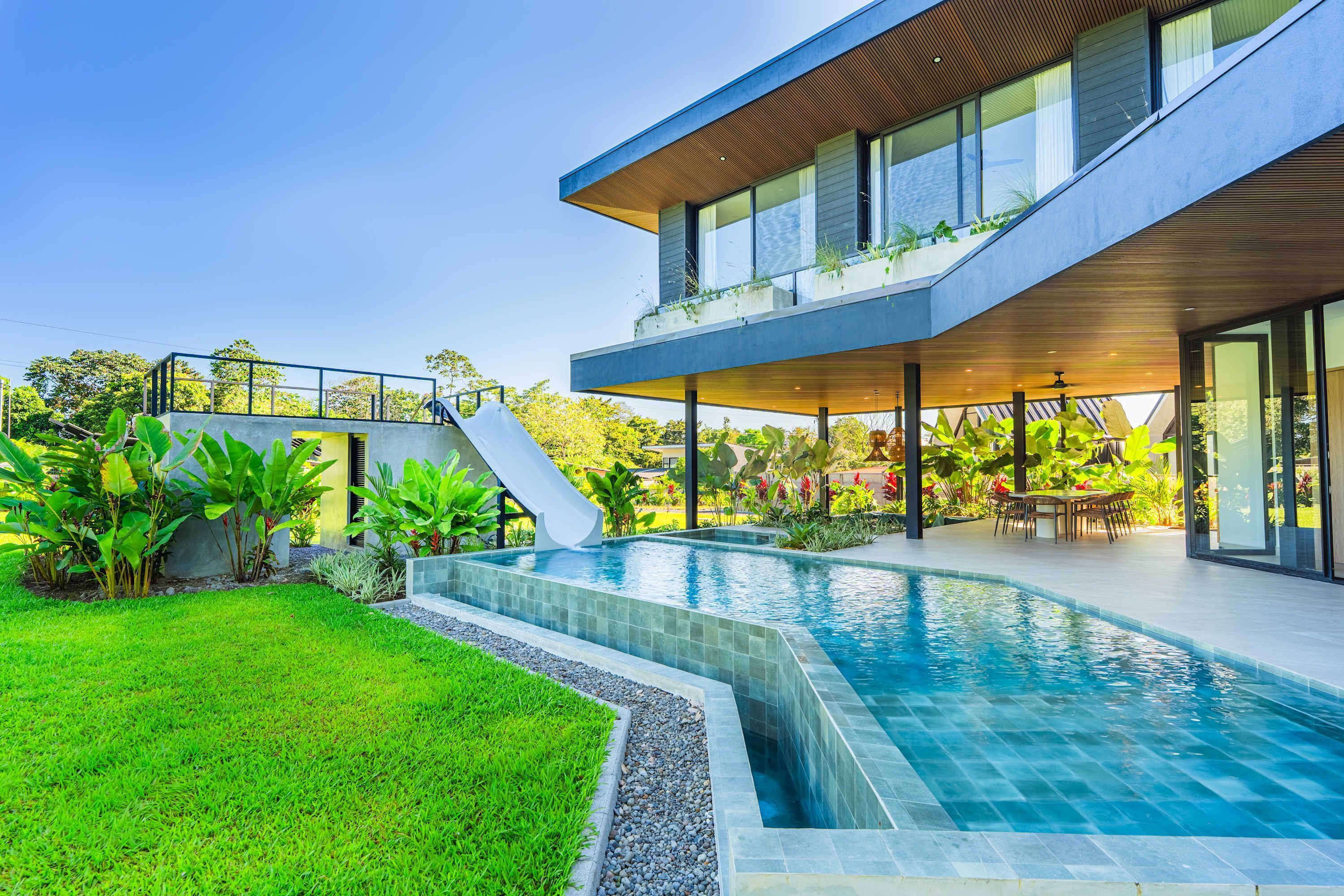 Una casa moderna de dos pisos con piscina, mobiliario exterior y abundante vegetación.