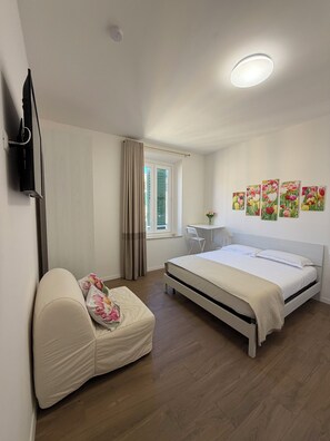Een slaapkamer met een bed, een bureau, een stoel, een televisie en een raam met gordijnen.