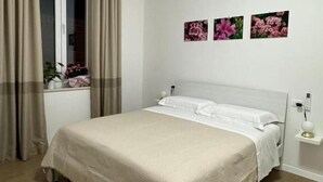 A bedroom with a bed, curtains, a nightstand, and wall art. - LINDA affittacamere (ANCONA)