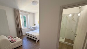 A bedroom with a bed, a bedside table, a chair, a small table with flowers, and a shower area. - LINDA affittacamere (ANCONA)