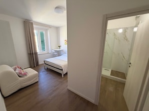 Een slaapkamer met een bed, nachtkastje, stoel, een klein tafeltje met bloemen en een douchegedeelte.