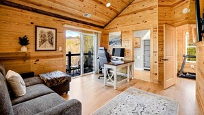 Ein gemütliches Blockhauskamin-Interieur mit Holzbalkendecke, einer Couch, einem Schreibtisch mit Computer und einer Glastür zum Balkon.