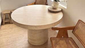 Mesa redonda com base trançada, rodeada por cadeiras de madeira com encostos de palha, uma orquídea em vaso sobre um apoio de palha e vista para uma área residencial através de grandes janelas.