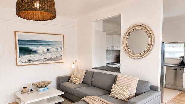 Uma sala de estar moderna com um sofá cinzento, uma mesa de centro, um quadro grande emoldurado com vista para o oceano, uma luminária de vime e uma cozinha visível através de uma porta aberta.