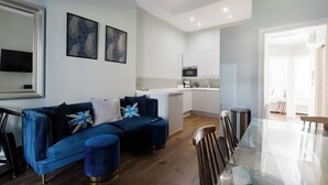 A modern living room with a blue sofa, a glass dining table, and a kitchen in the background. - Stylish 3BDR Flat 10 Minutes from Portobello Road (Greater London)