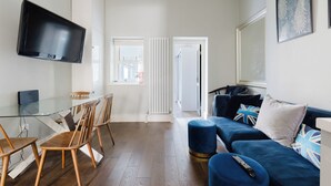 A modern living room with a glass dining table, wooden chairs, a blue sofa with decorative pillows, and a mounted television. - Stylish 3BDR Flat 10 Minutes from Portobello Road (Greater London)
