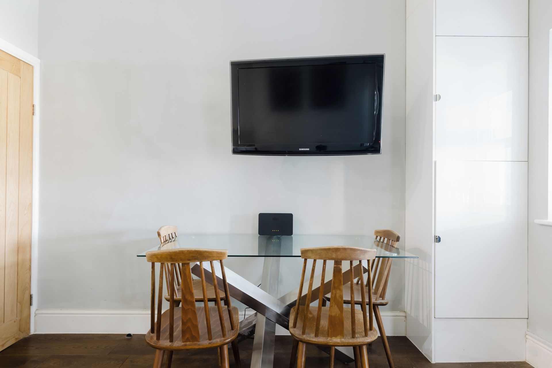 Una habitación con una mesa de vidrio, dos sillas de madera, un televisor de pantalla plana montado en la pared y un armario blanco.