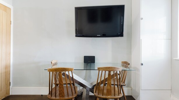 A room with a glass-top table, two wooden chairs, a flat-screen TV mounted on the wall, and a white cabinet. - Stylish 3BDR Flat 10 Minutes from Portobello Road (Greater London)