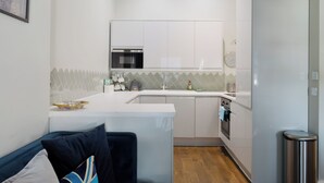 A compact kitchen with a microwave, oven, and sink. There is a small dining area with a couch and a trash bin. - Stylish 3BDR Flat 10 Minutes from Portobello Road (Greater London)