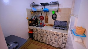 A compact kitchen with a tiled backsplash, a sink, a countertop with various kitchen items, and a small trash bin. - Jurassic Surf House Anza (Agadir)