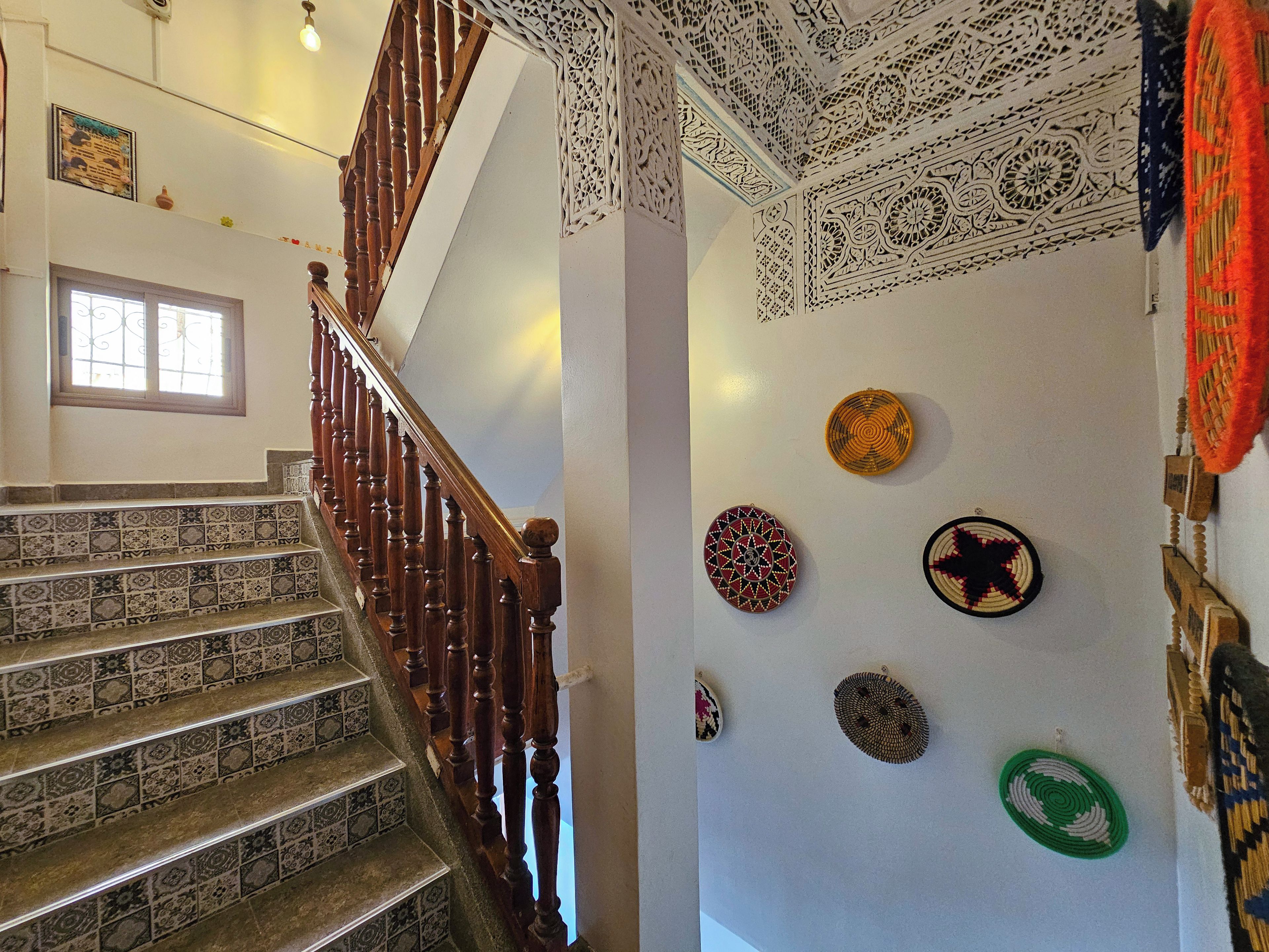 A staircase with patterned tiles and a wooden handrail, leading to a room with a white wall adorned with various decorative plates.