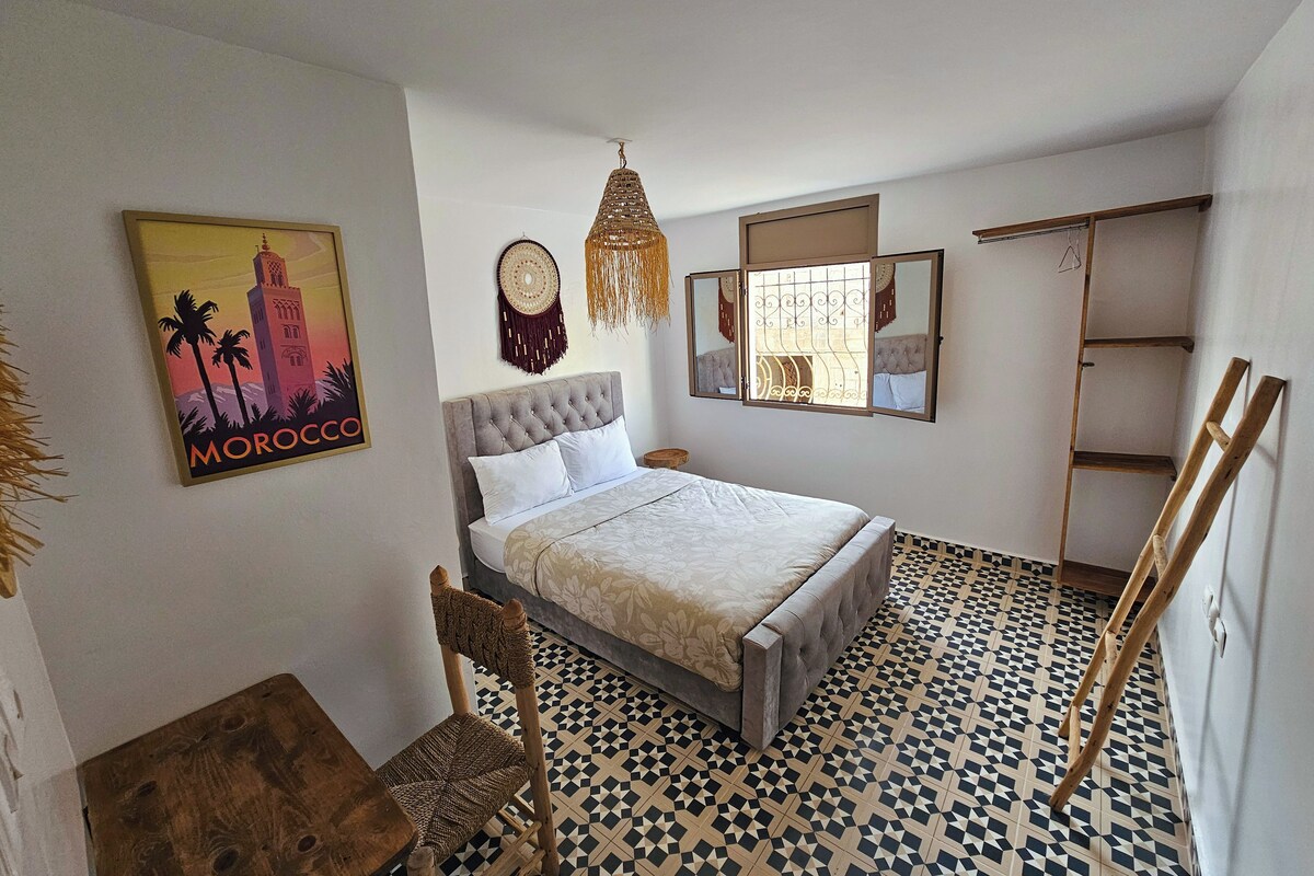 Un dormitorio con cama, una silla, un espejo montado en la pared, un adorno en la pared y un cuadro enmarcado.
