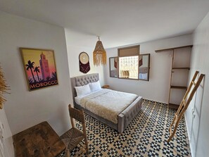 Un dormitorio con cama, una silla, un espejo montado en la pared, un adorno en la pared y un cuadro enmarcado.