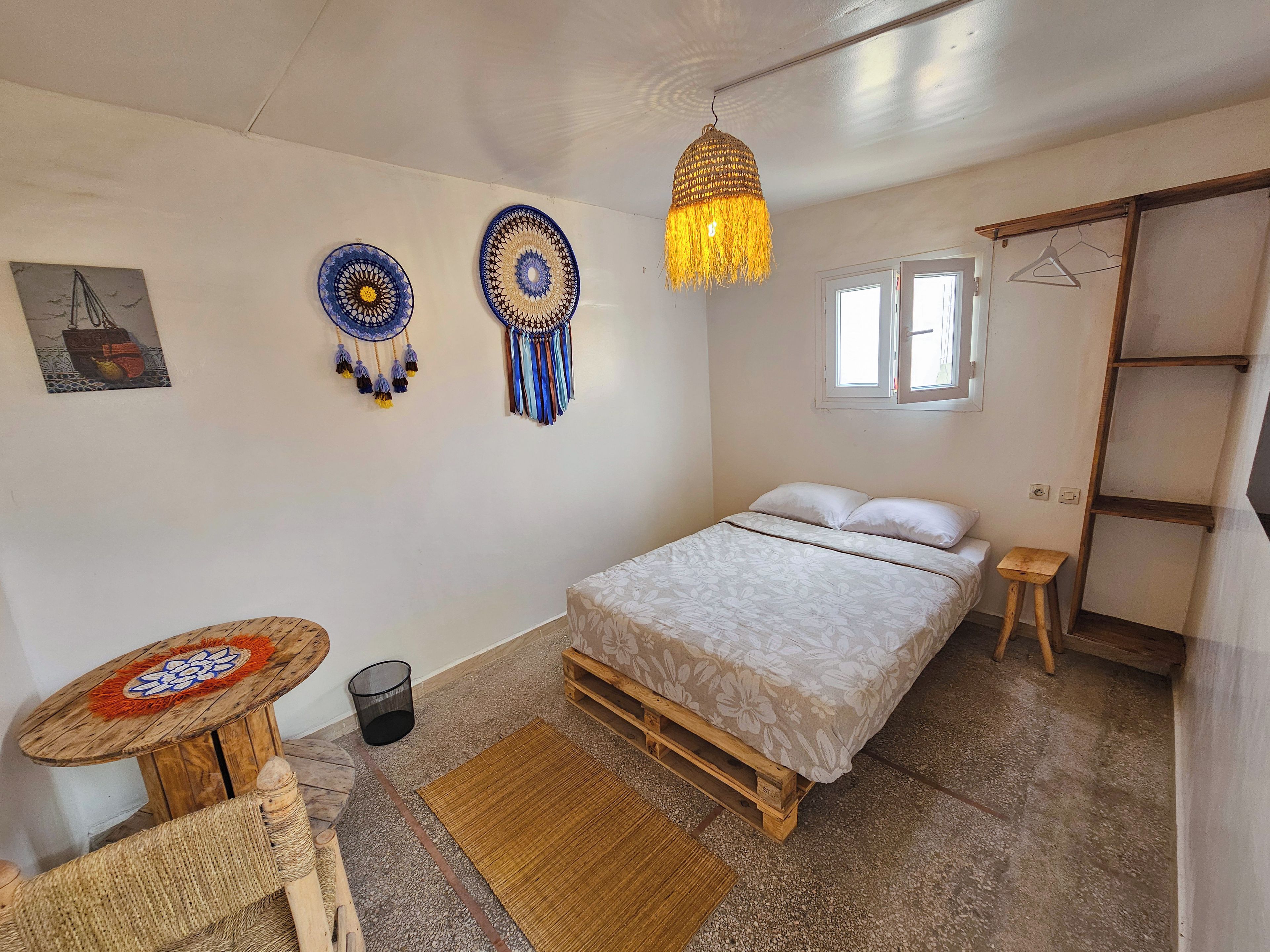 Un dormitorio con cama, lámpara colgante, mesita, silla y estante de pared.