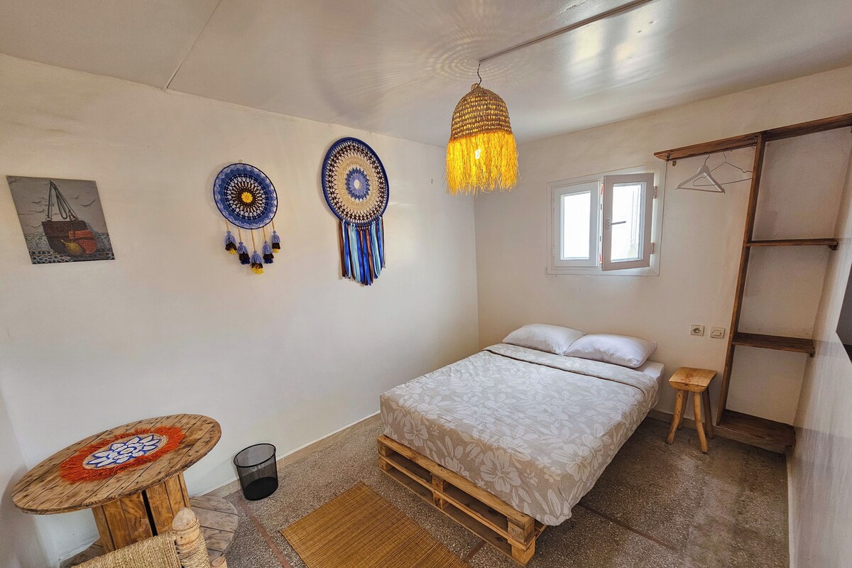 Un dormitorio con cama, lámpara colgante, mesita, silla y estante de pared.