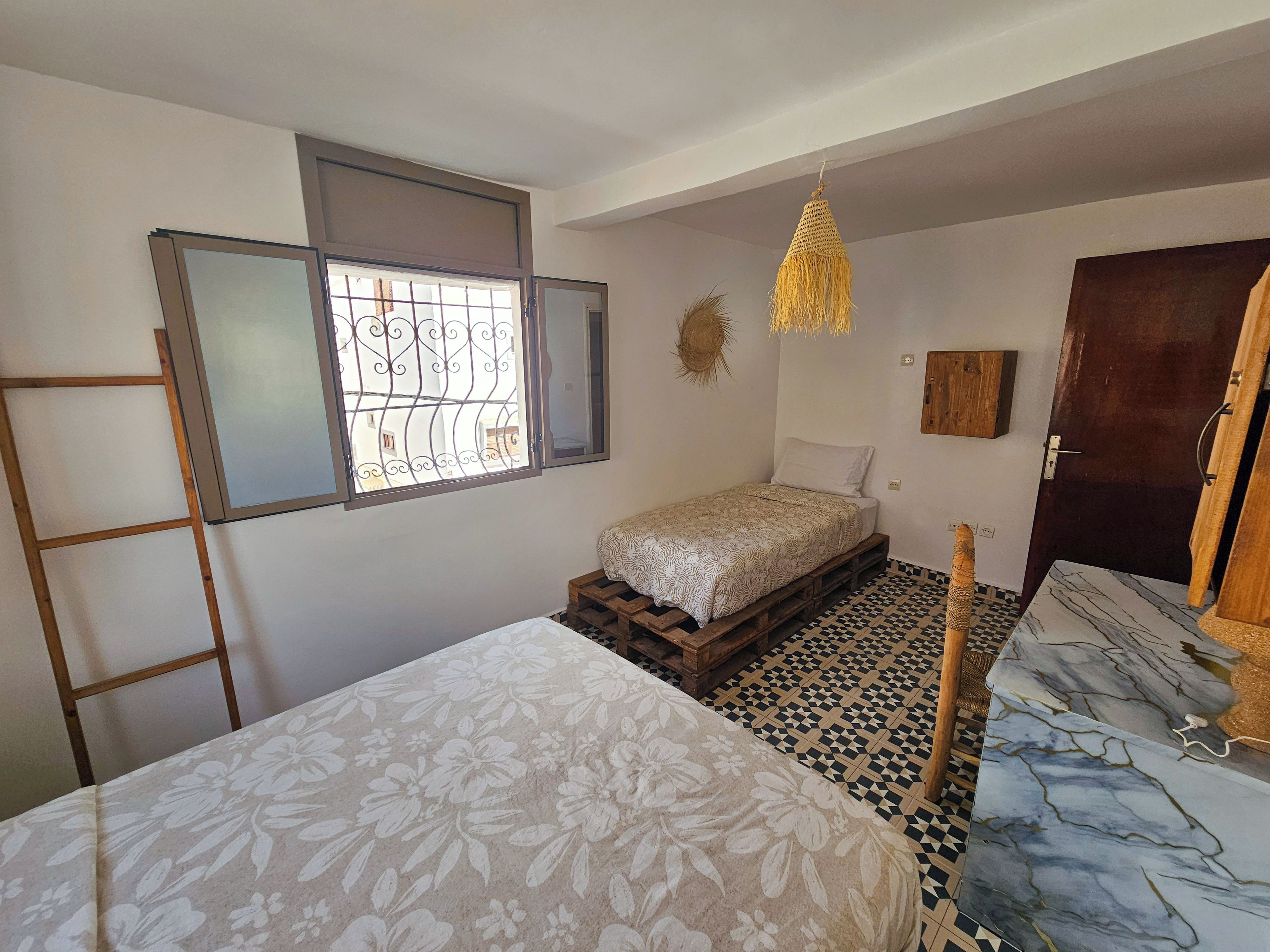 A room with a bed, a window with bars, a hanging light fixture, and a door.