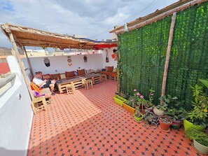 A rooftop patio with a wooden table and benches, a person seated, and potted plants.